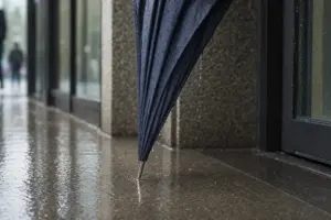 Punta metallica di un ombrello chiuso appoggiata sul marciapiede bagnato, con gocce d’acqua che scendono