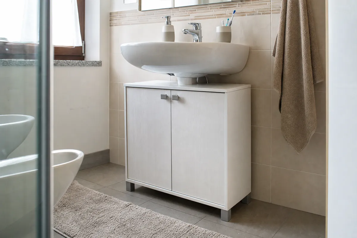 Mobile sotto lavabo bianco a due ante sotto un lavandino in ceramica, in bagno con piastrelle beige e asciugamano