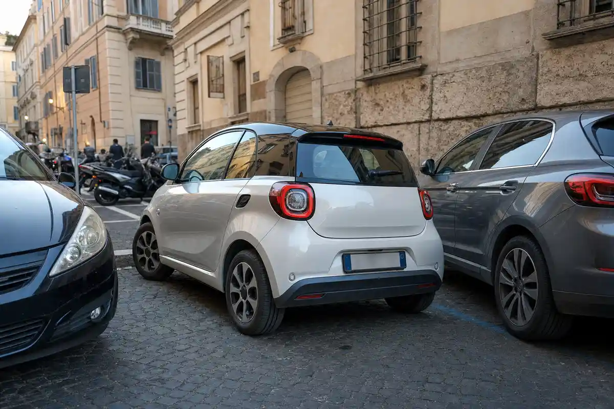 Citycar bianca a due porte parcheggiata stretta tra due auto su strada in sampietrini, con scooter sullo sfondo