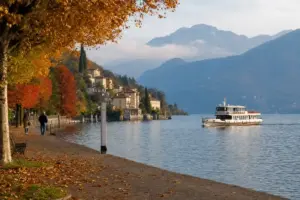 Battello sul lago con passeggiata alberata d’autunno, foglie a terra e due persone che camminano, montagne sullo sfondo
