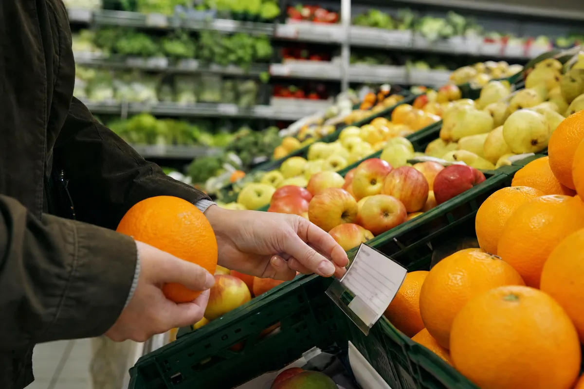 Mani di un cliente che tiene un’arancia e controlla un cartellino di origine nel reparto ortofrutta del supermercato