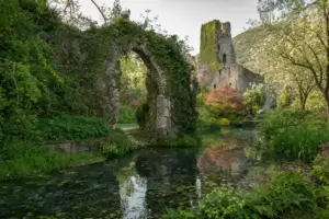 Rovine in pietra con arco coperto d’edera e torre medievale affacciate su uno stagno limpido tra fiori e alberi
