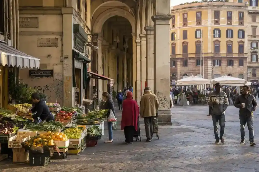 Bancarella di frutta e verdura sotto i portici all’Esquilino, con passanti e tavolini all’aperto in piazza