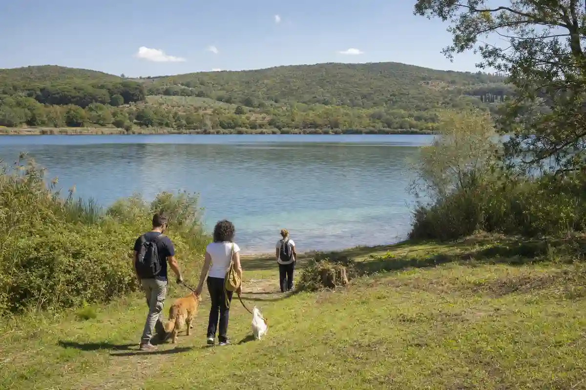 Persone con due cani al guinzaglio scendono lungo un sentiero erboso verso un lago limpido tra colline verdi