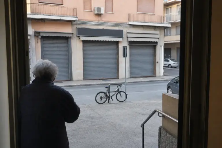 Persona anziana vista di spalle osserva una fila di negozi con saracinesche abbassate, con bici legata a un palo