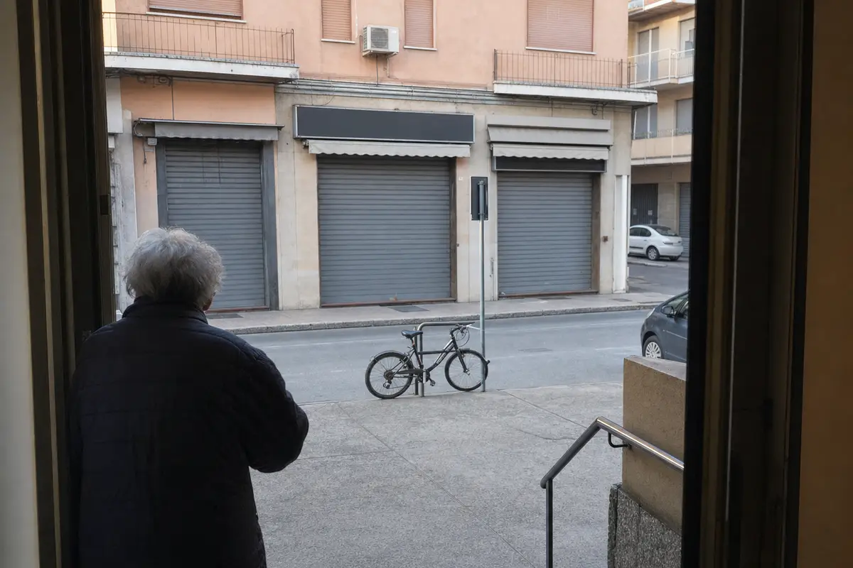 Persona anziana vista di spalle osserva una fila di negozi con saracinesche abbassate, con bici legata a un palo