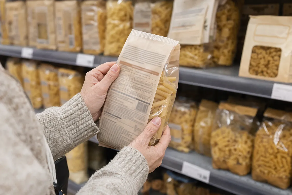 Mani di una persona che legge l’etichetta sul retro di un pacco di pasta in corsia supermercato