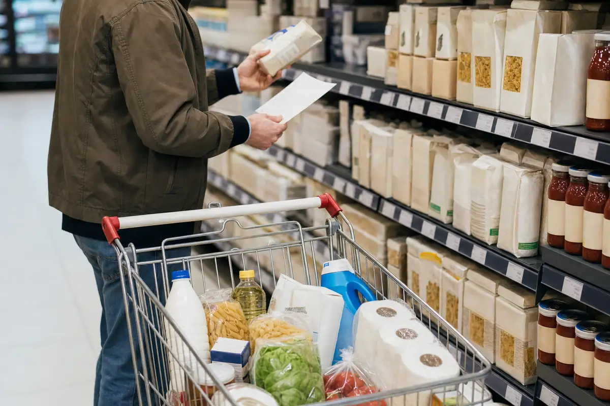 Cliente in corsia supermercato confronta un prodotto con lista della spesa accanto a un carrello con alimenti e carta