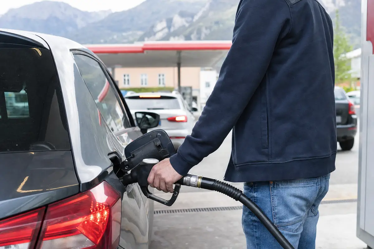 Persona fa rifornimento a un’auto grigia con la pistola della benzina in un distributore, con altre auto sullo sfondo
