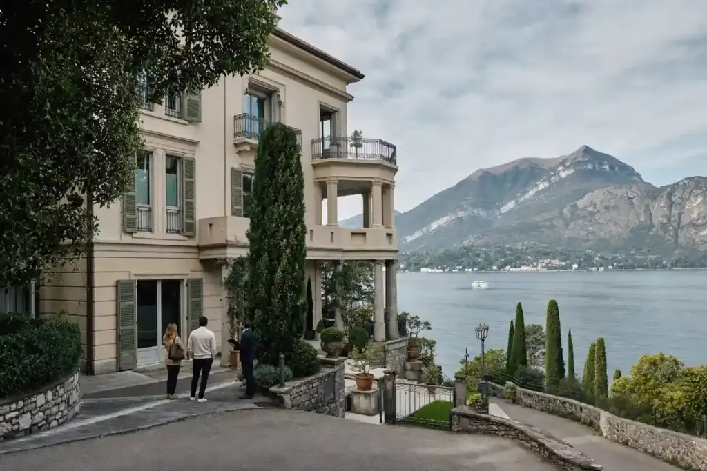 Villa elegante sul lago con balconi, cipressi e tre persone che parlano nel vialetto, montagne sullo sfondo