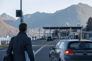 Pendolare con cartelle e borsa vicino a un valico di confine, con auto in coda e montagne sullo sfondo