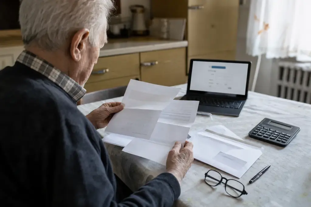 Anziano al tavolo di casa che controlla documenti e buste con laptop aperto e calcolatrice accanto