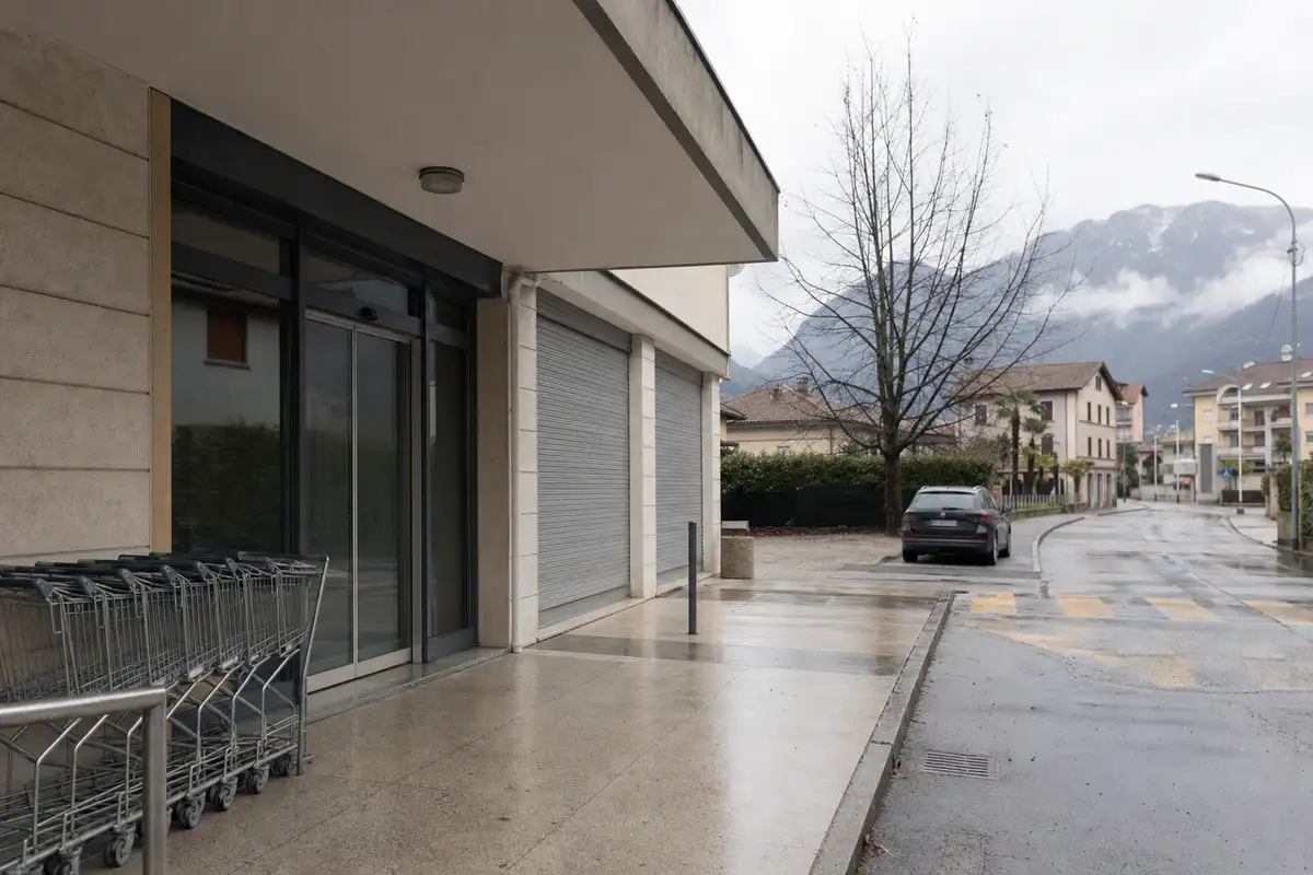 Ingresso di un supermercato chiuso con serrande abbassate e carrelli vuoti in una strada bagnata, con montagne sullo sfondo