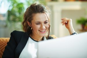 happy elegant female in white blouse and black jacket at modern home in sunny day doing research on a laptop while sitting on couch.