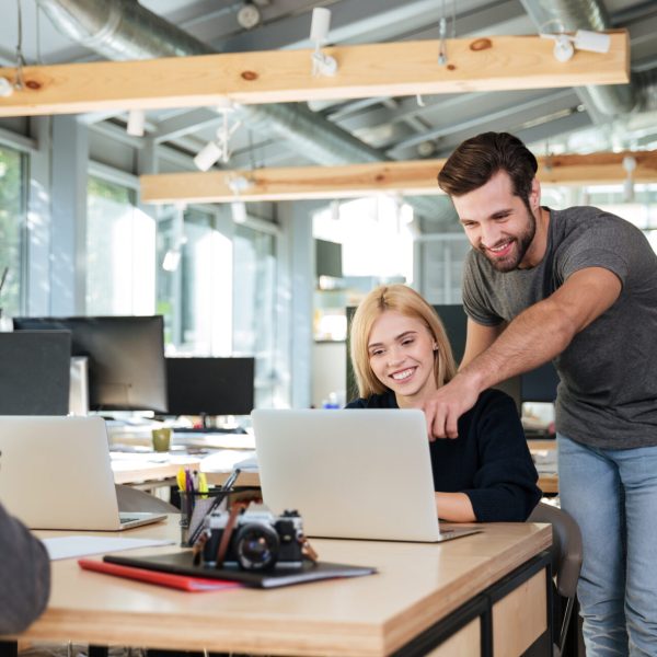 Picture of cheerful young colleagues sitting in office coworking using laptop computers. Looking aside.