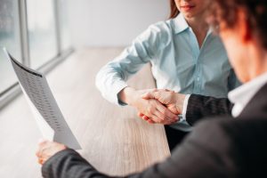 Unrecognizable businesswoman holding document and shaking hand of business partner after signing of contract. They sitting at table. Negotiation concept