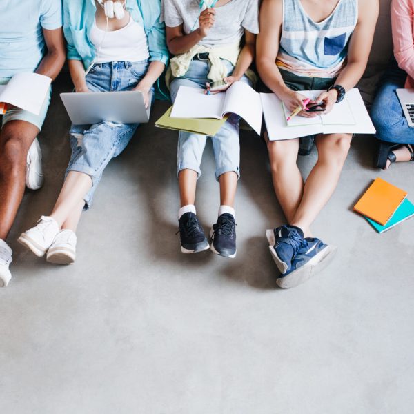 Overhead portrait of young people with laptops and smartphones, sitting together on the floor. Students writing lectures holding textbooks on their knees.