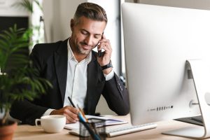 Image of handsome businessman 30s wearing suit talking on cell phone while working on computer in office